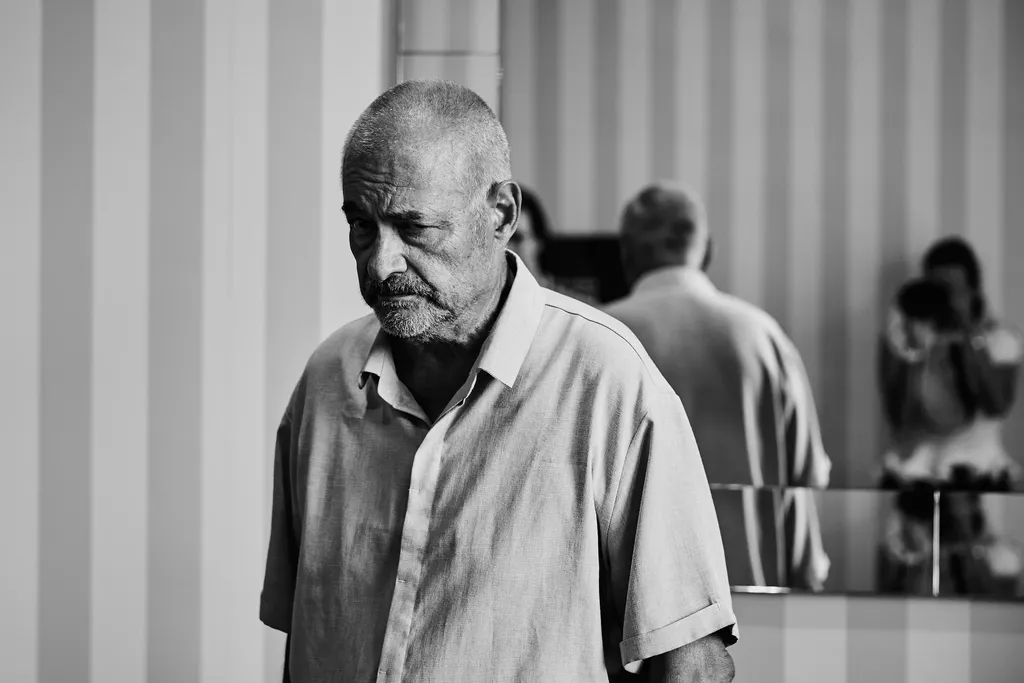An older man in a light shirt, head lowered, in front of a striped wall and a mirror reflecting someone taking a picture, black and white.