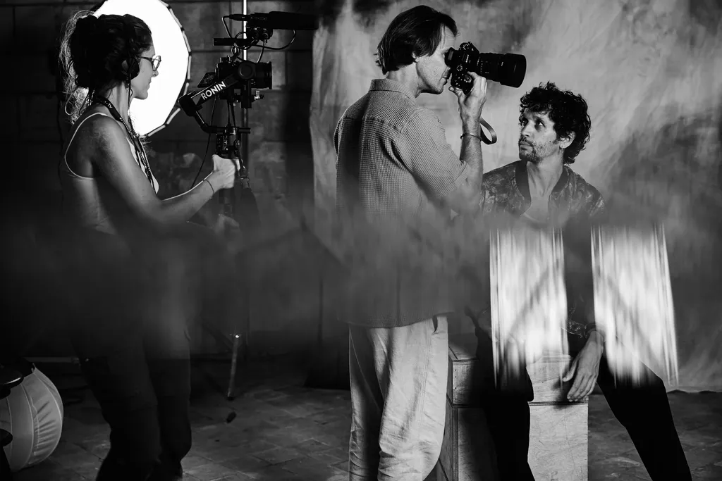A camerawoman beside a ring light while a man photographs another man up close, black and white.
