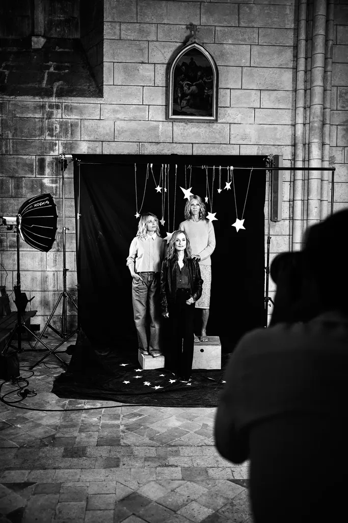 Three women pose on crates in front of a black backdrop hung with stars, a softbox on the left, in a stone hall, black and white.
