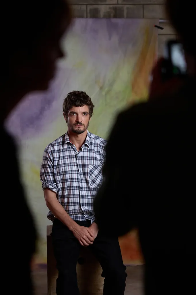 A man in a checked shirt seated before a painted backdrop, framed between two dark silhouettes, colour.