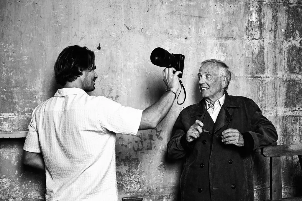 A man holds a camera out toward an older man in a dark coat against a worn wall, black and white.