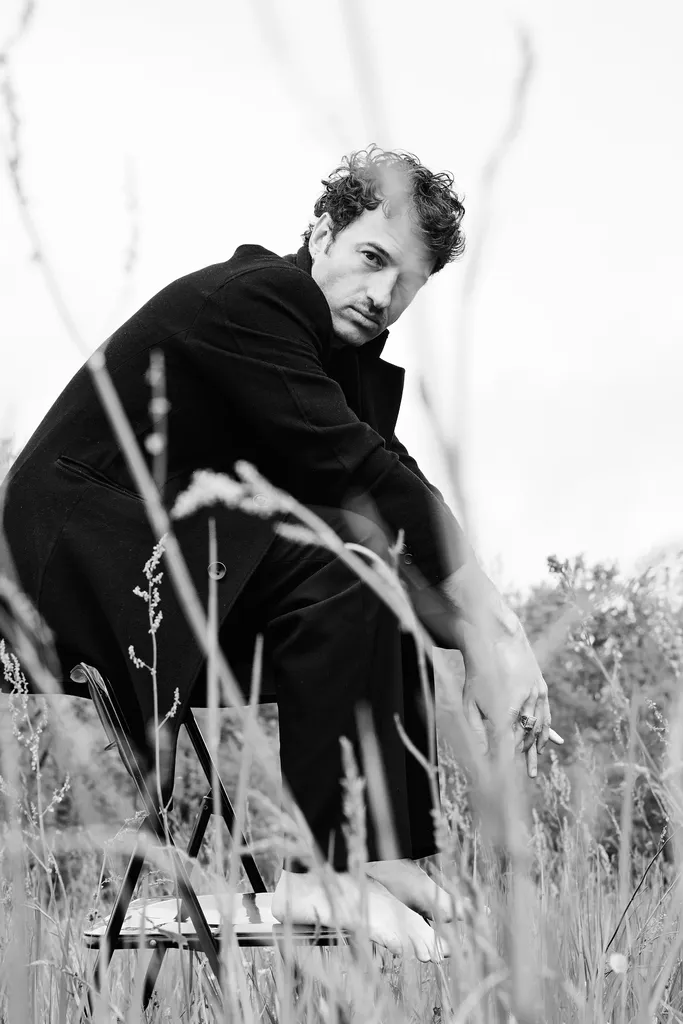 A man in a black coat crouched on a folding chair, barefoot in tall grass, black and white.
