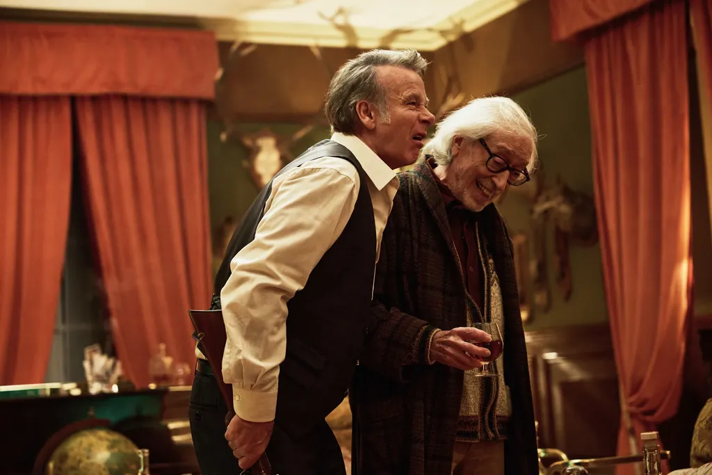 Two men laughing in a period drawing room with red curtains, one holding a glass, colour.