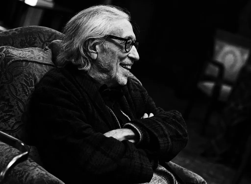 An older man in glasses smiling, arms folded in a patterned armchair, low light, black and white.