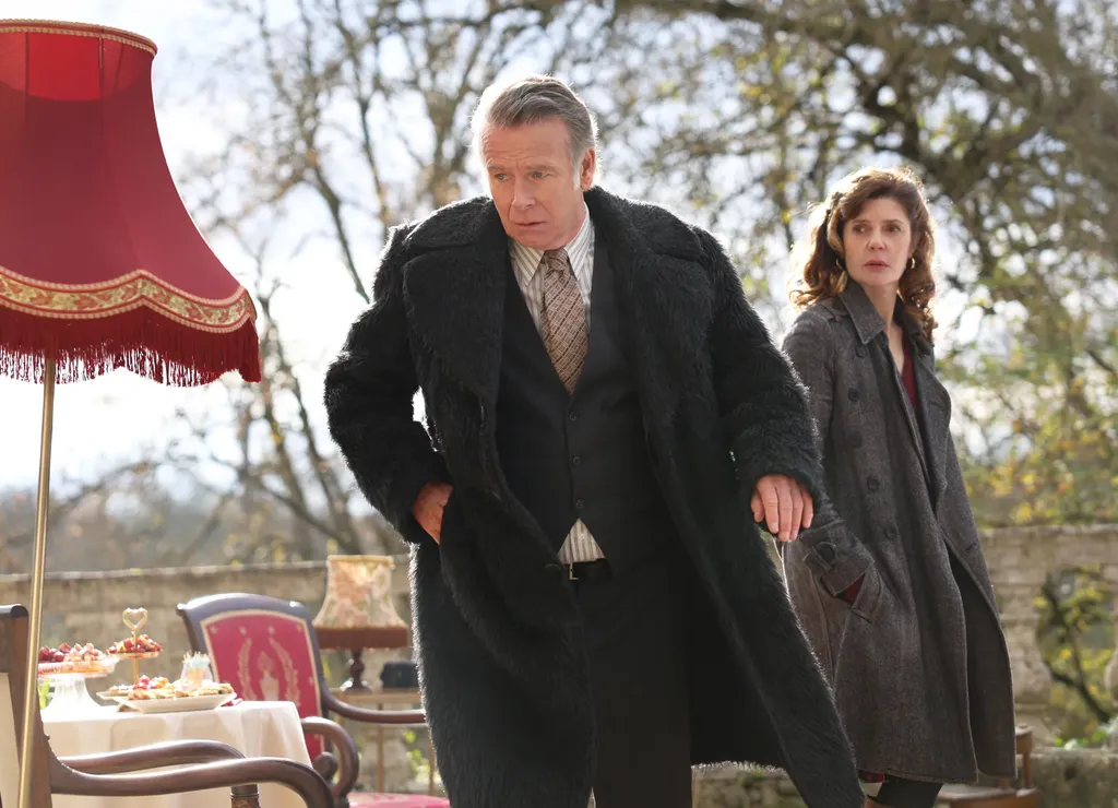 A man in a dark coat and a woman in a grey coat in a garden, beside a red fringed lampshade and a laid table, colour.