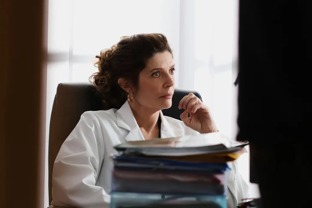 A woman in a white coat seated at a desk behind a stack of files, window light, colour.