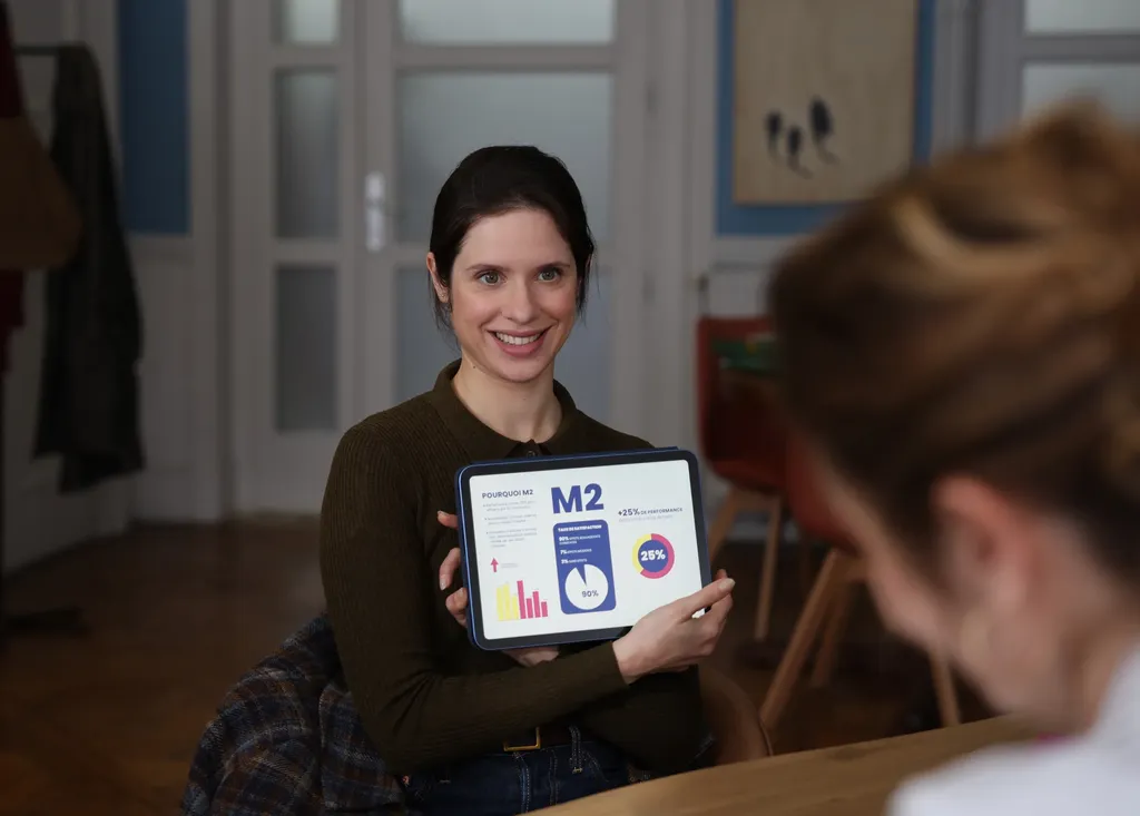 A smiling woman holds up a tablet, seen over another person’s shoulder, in a bright flat, colour.
