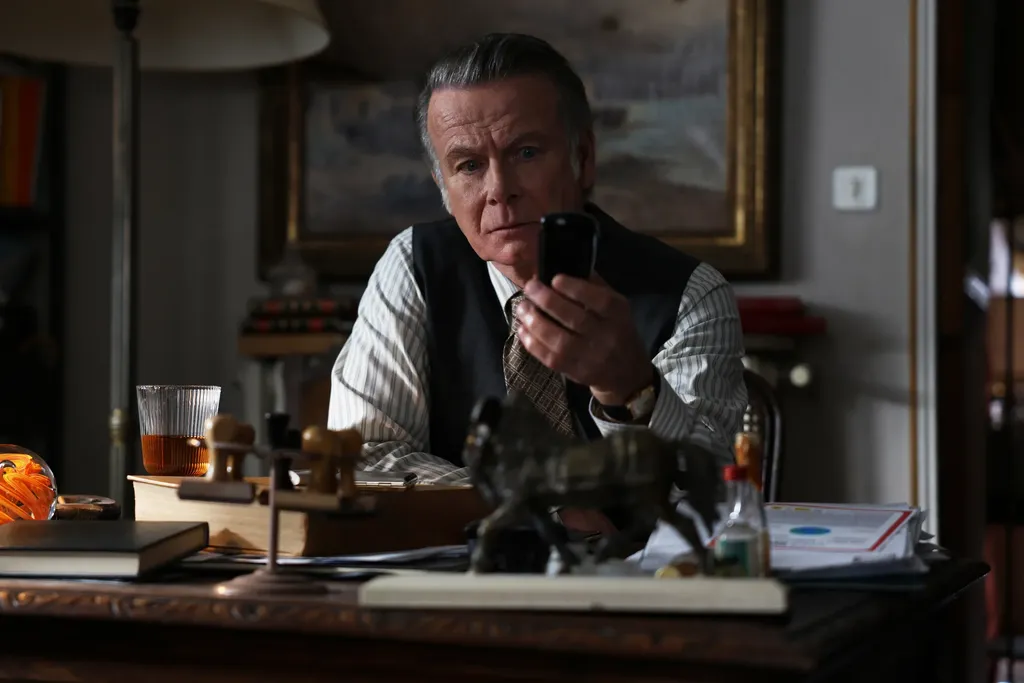 A man in a waistcoat at a cluttered desk, phone in hand, a horse figurine in front of him, colour.