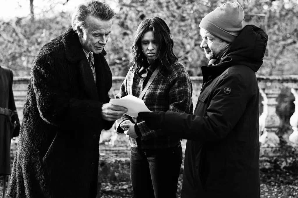 Three people outdoors reading a sheet together: a man in a fur coat, a young woman and a man in a beanie, black and white.