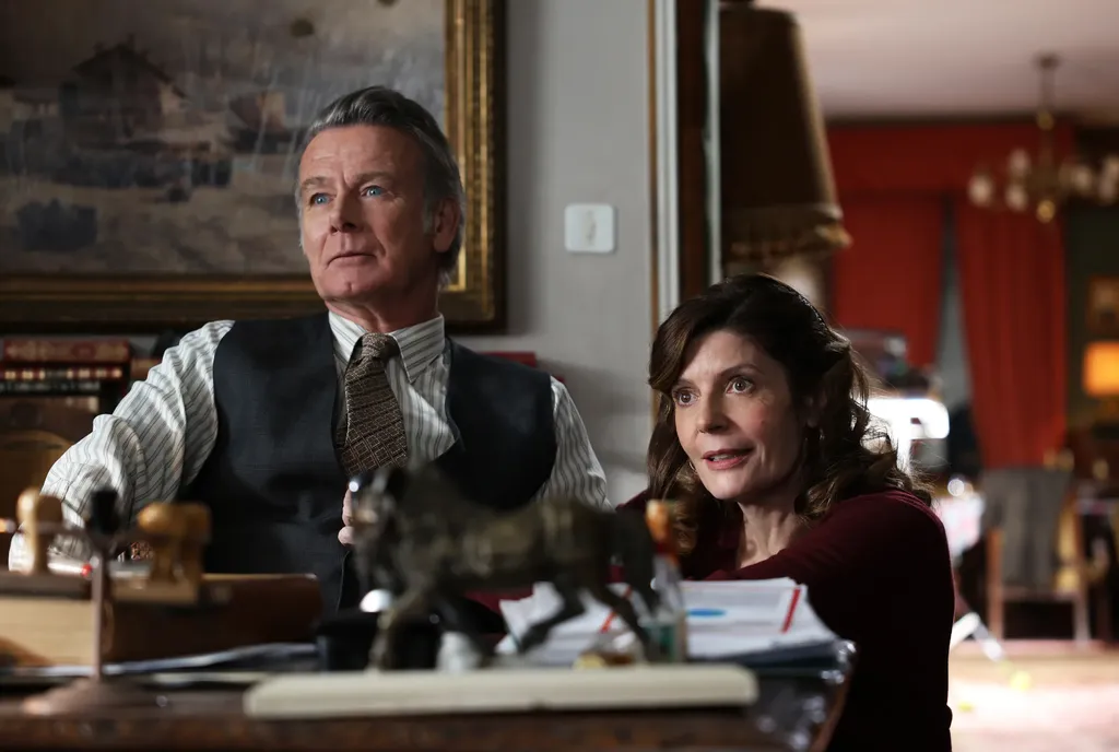 A man in a waistcoat and tie seated at a desk, a woman in a red jumper leaning beside him, red curtains behind, colour.