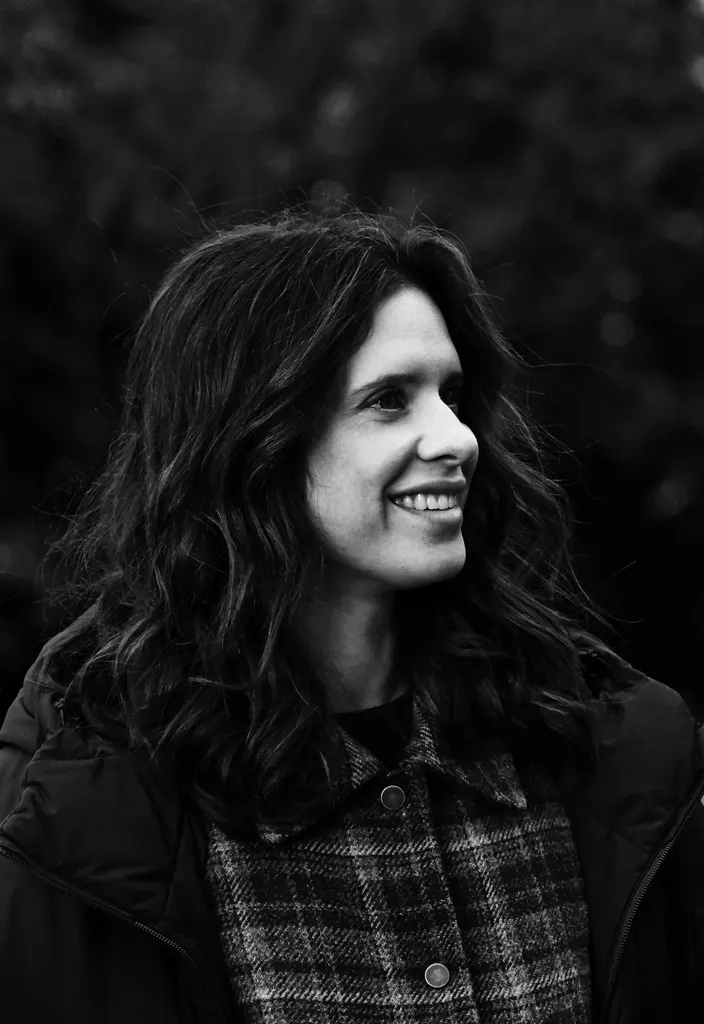 Portrait of a woman with long dark hair smiling and looking aside, dark foliage behind, black and white.