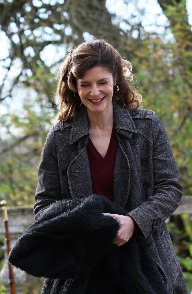 A smiling woman in a grey coat carrying a black fur coat over her arm, autumn trees behind her, colour.