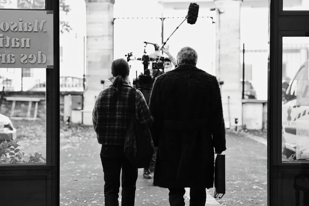 Seen through a glass door, two people walk toward a crew and a boom pole under a stone gateway, black and white.