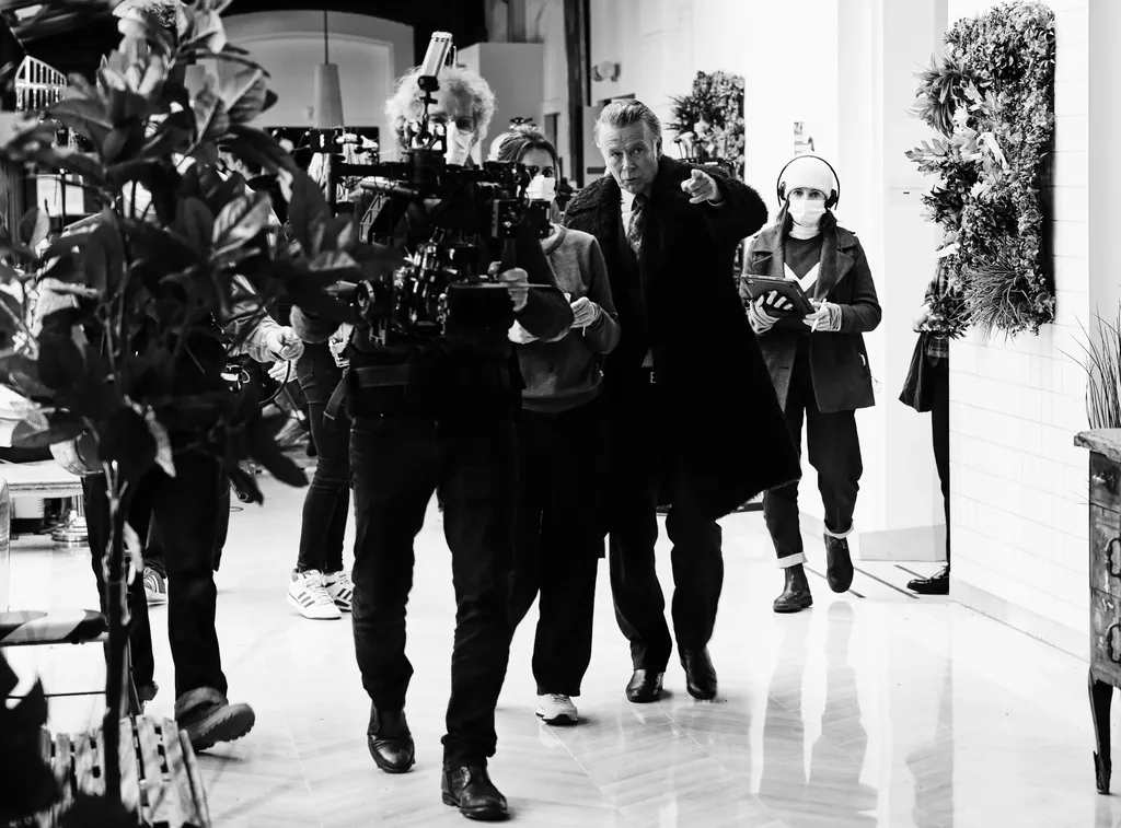 A crew moving through a white tiled hall, an operator with the camera on his shoulder, a man pointing, a woman with headphones and a tablet, black and white.