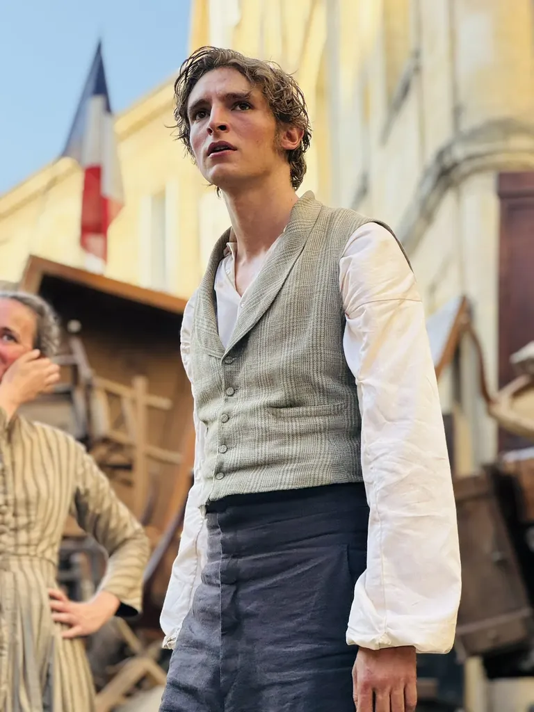 A young man in period costume, waistcoat and white shirt, in a street with a tricolour flag and wooden debris, colour.