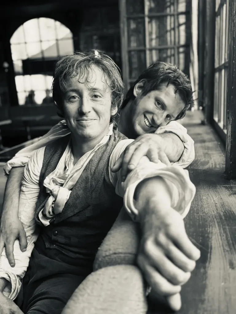 Two young men in period costume smiling, one leaning on the other by a window, black and white.