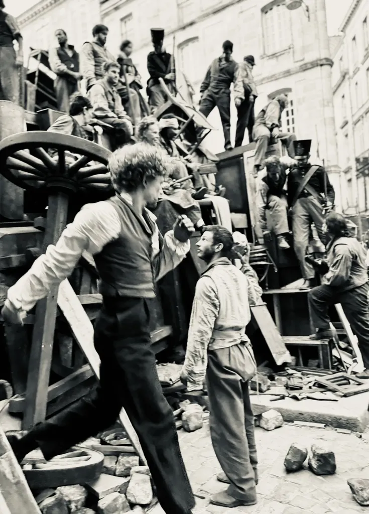 Extras in period costume climb a barricade of furniture and cartwheels in a street, black and white.