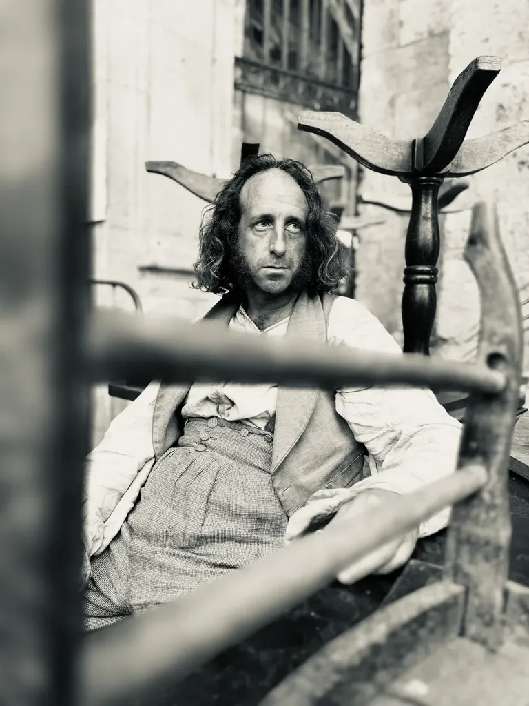 A long-haired man in period clothes seated behind wooden rails and a coat stand, black and white.