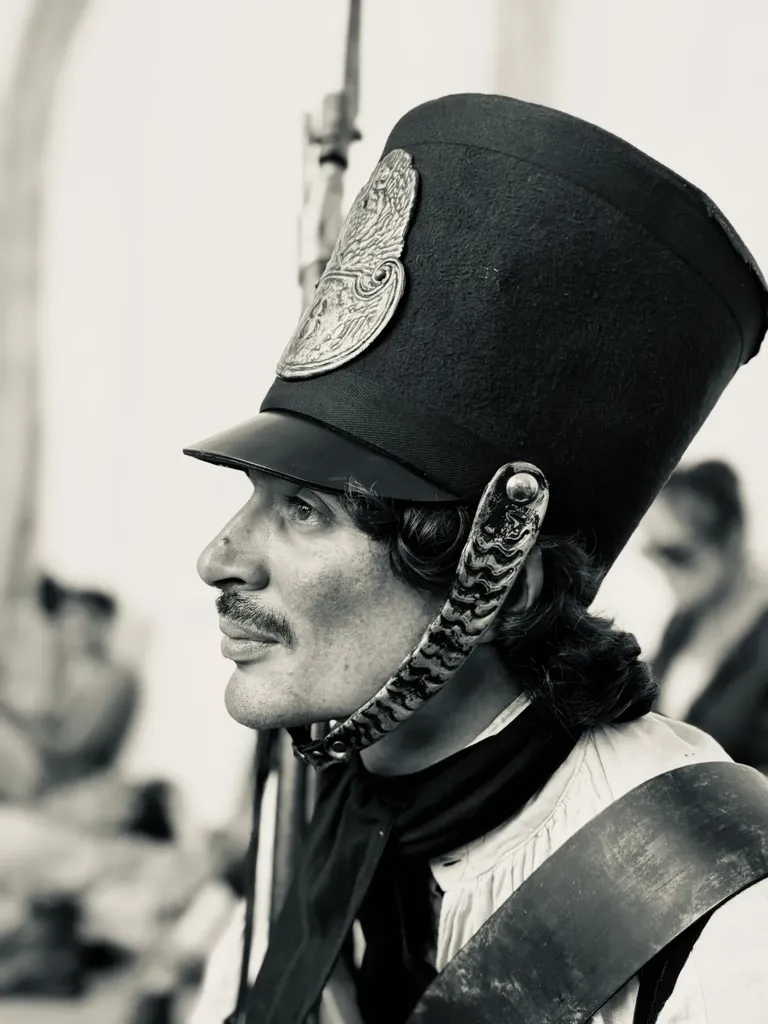A soldier in profile wearing a tall shako with a metal plate and chin strap, black and white.