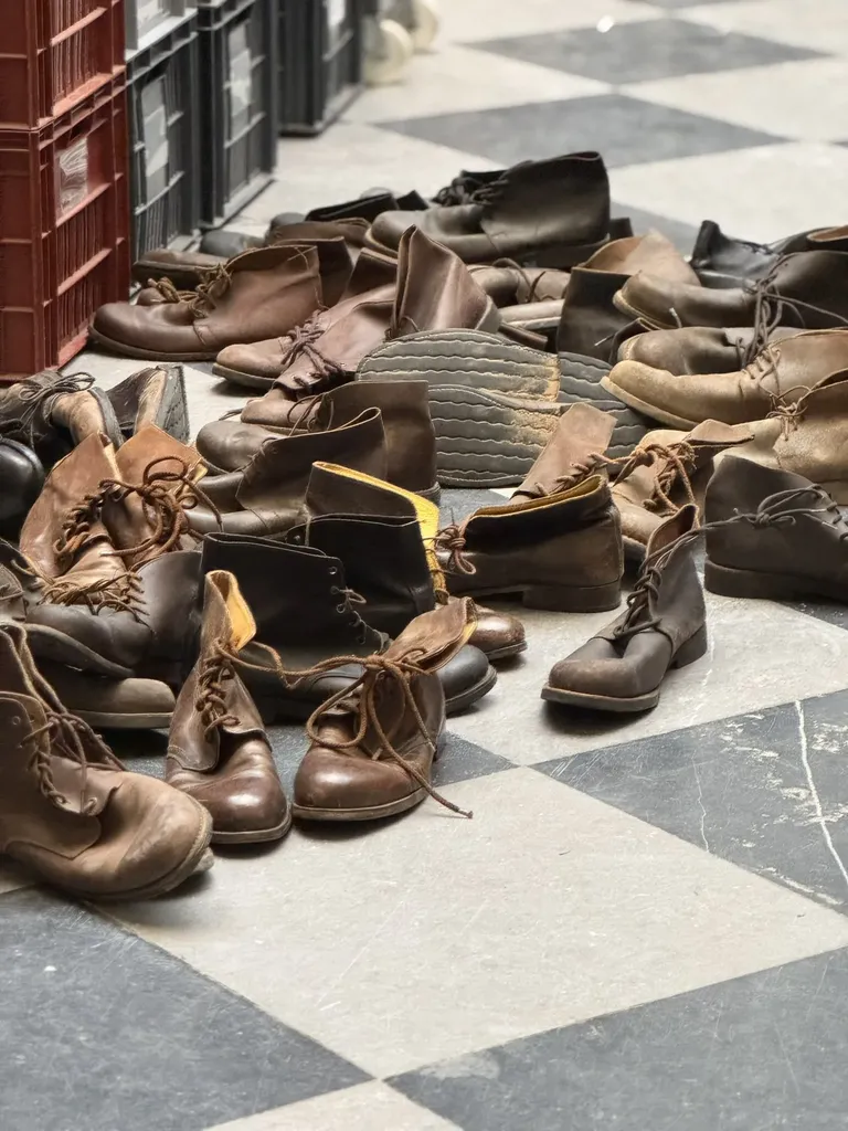 A heap of worn leather boots on a black and white chequered floor, next to red crates, colour.