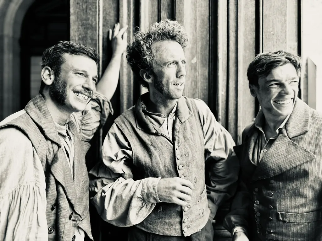 Three men in period costume laughing in front of a wooden door, black and white.