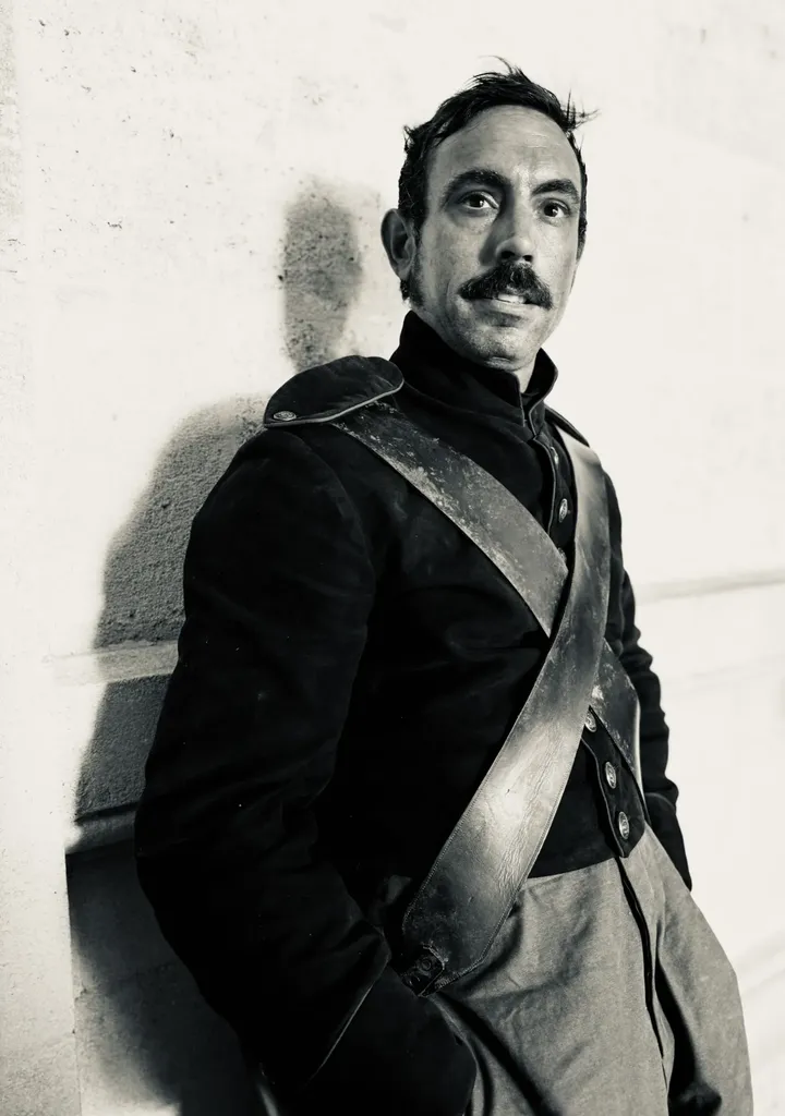 A man with a moustache in a period uniform with a cross belt, leaning on a pale wall, black and white.