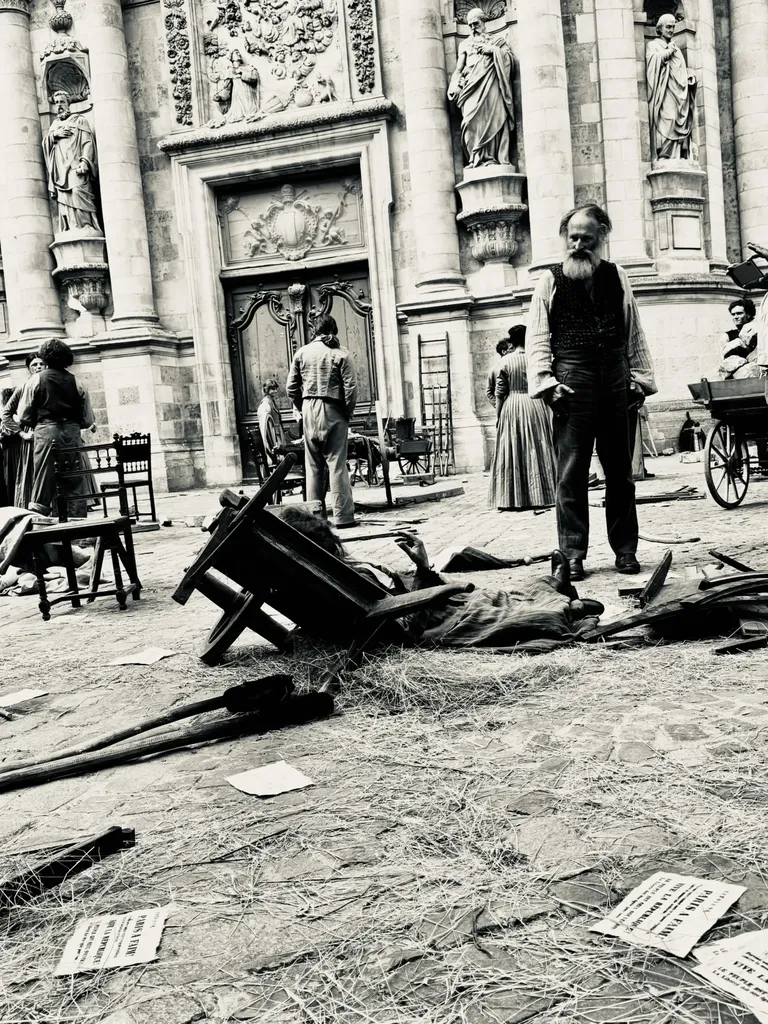 A square before a carved church front, chairs overturned, straw and papers on the ground, extras in costume, black and white.