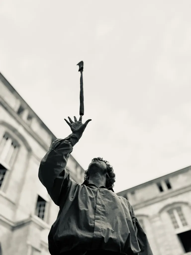From below, a man in costume reaches toward a stick thrown in the air between two building fronts, black and white.