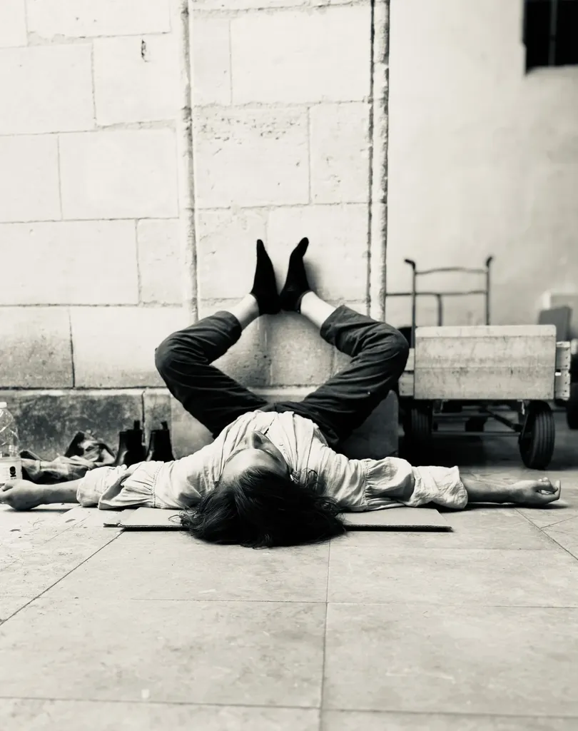 An actor lying on his back at the foot of a stone wall, legs raised against it, a cart behind, black and white.