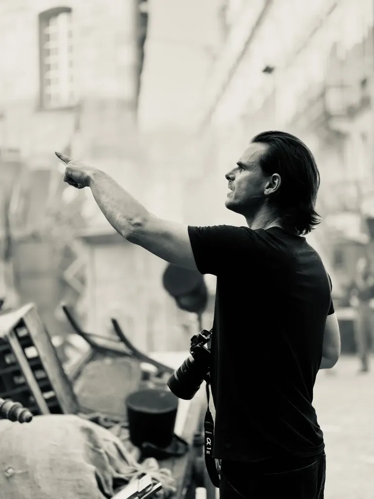 A man in a black T-shirt with a camera on a strap points at something on a street set, black and white.