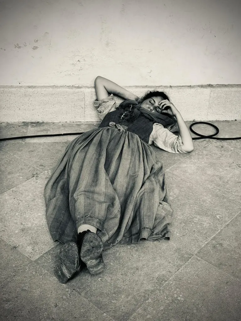 A woman in a long period dress lying at the foot of a wall on a stone floor, hand on her forehead, a cable beside her, black and white.