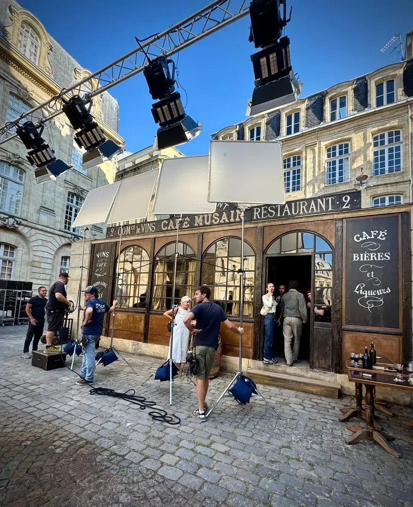 A café set on a cobbled square, a truss of lamps and diffusion frames above the crew, blue sky, colour.
