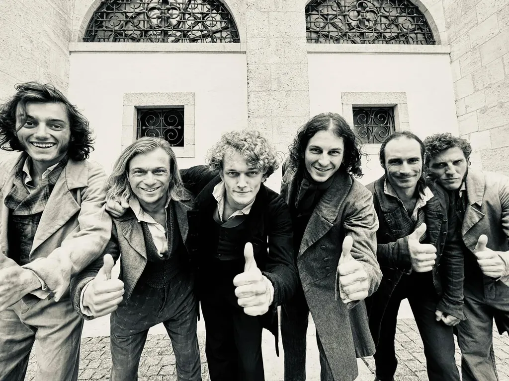 Six men in period costume lean toward the camera, thumbs up, in front of a stone building, black and white.