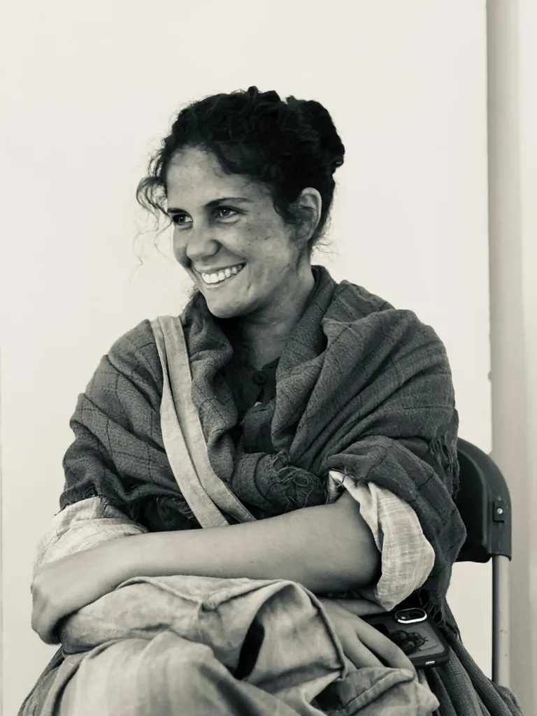 A smiling woman wrapped in a shawl, seated on a folding chair, arms folded, black and white.
