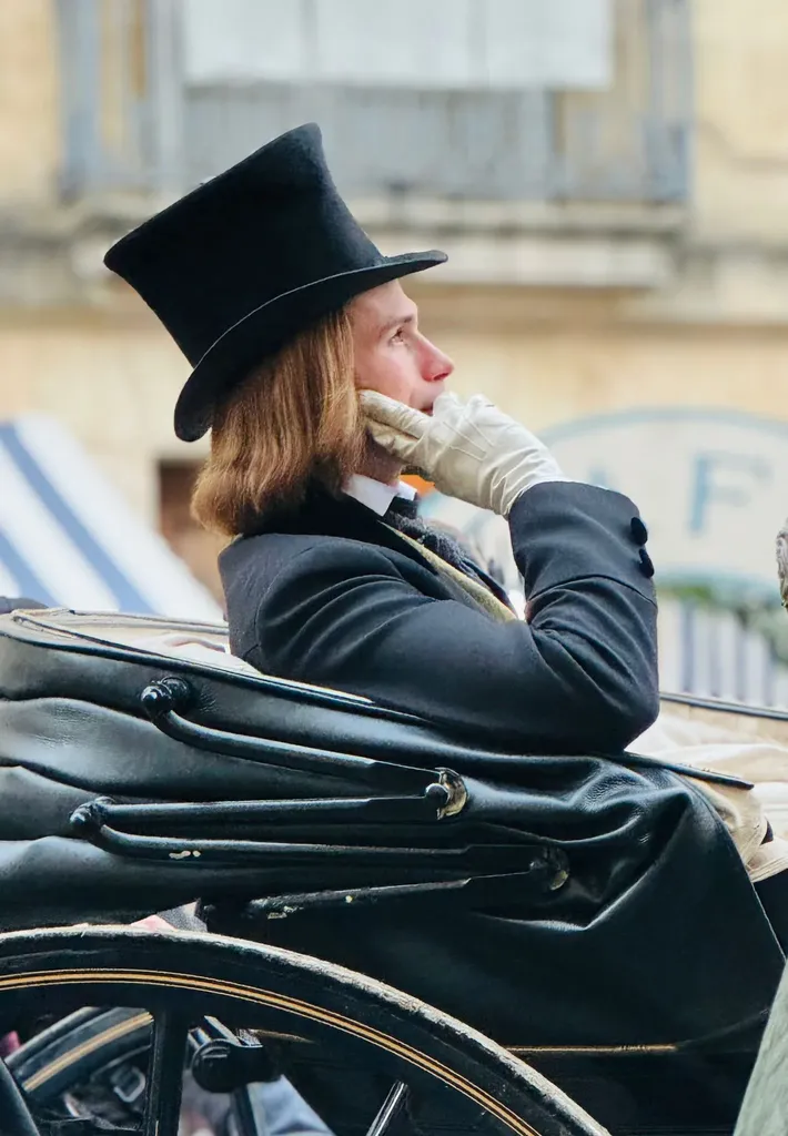 A long-haired young man in a top hat and pale gloves seated in a carriage, hand to his face, colour.