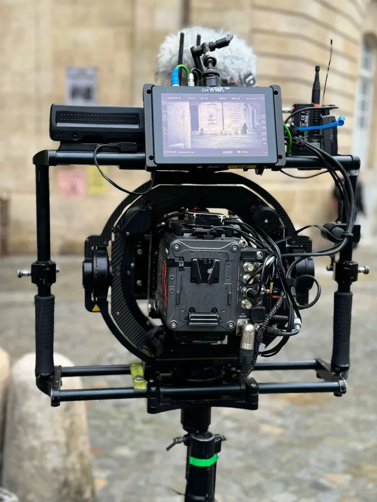 A cinema camera on a ring stabiliser, its monitor lit, on a cobbled square, colour.