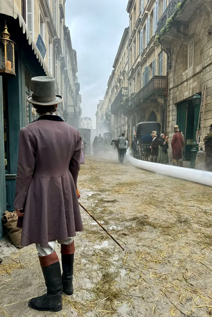 From behind, a man in a plum frock coat, top hat and boots walks down a street strewn with straw and smoke, colour.