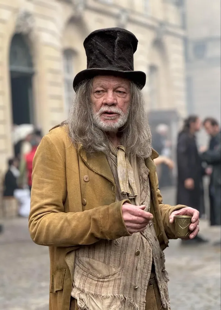An older man with long grey hair and a white beard, top hat and worn ochre coat, holding a cup, colour.