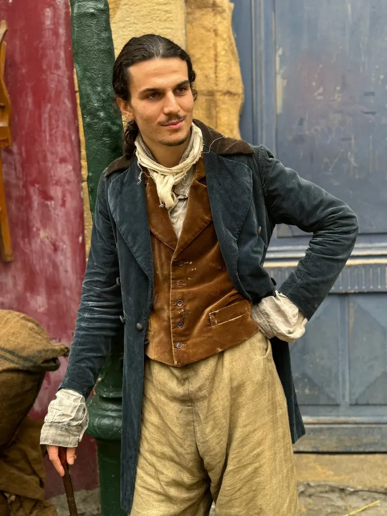 A young man in a velvet frock coat and brown waistcoat, hand on hip, before a red and blue wall, colour.