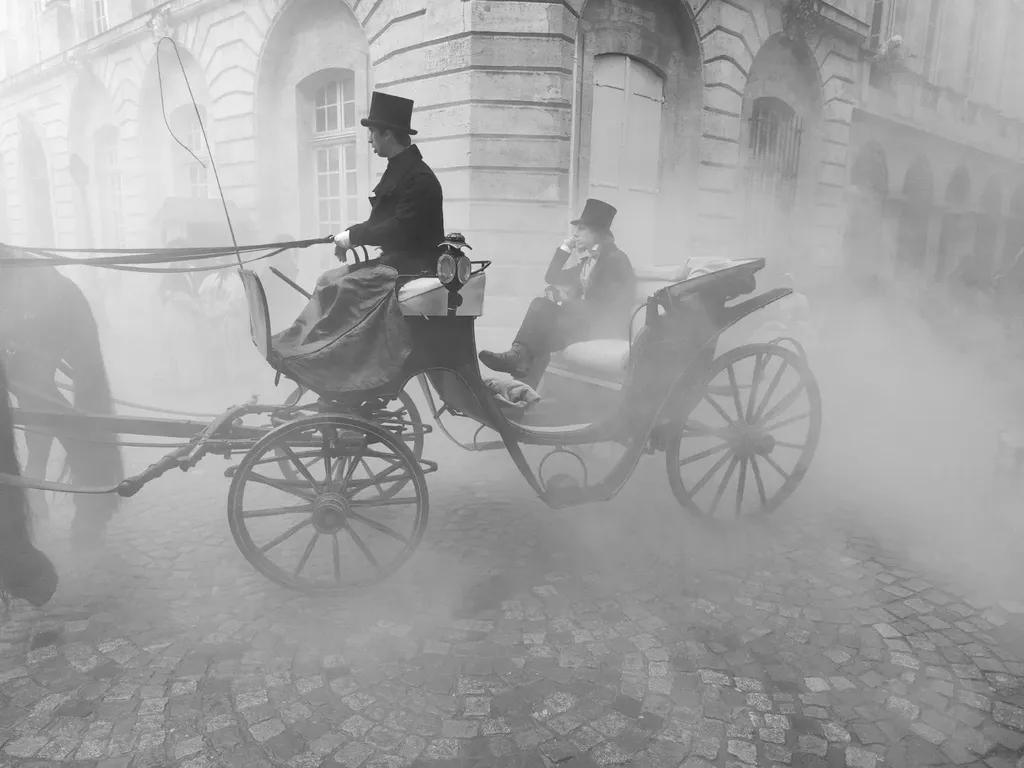 A carriage with a coachman and a passenger in top hats crosses a cobbled street thick with smoke, black and white.