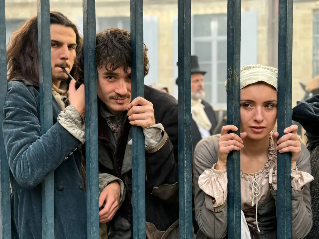 Three young people in period costume holding the bars of a blue gate, one smoking a pipe, colour.
