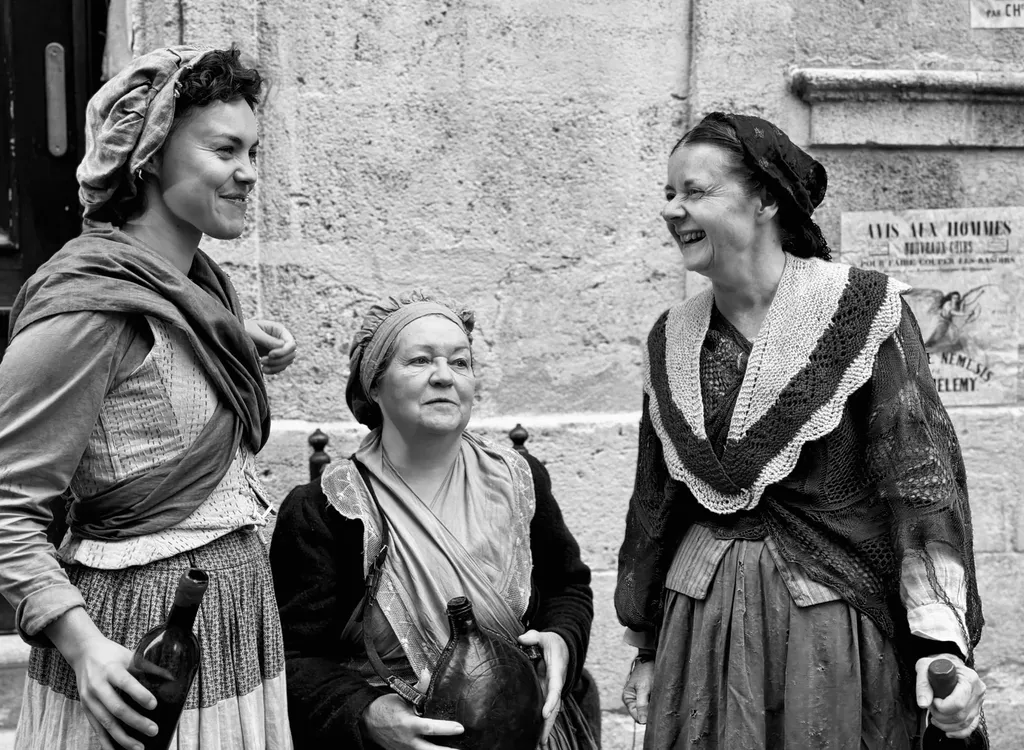 Three women in period dresses and headscarves laughing in front of a stone wall, black and white.