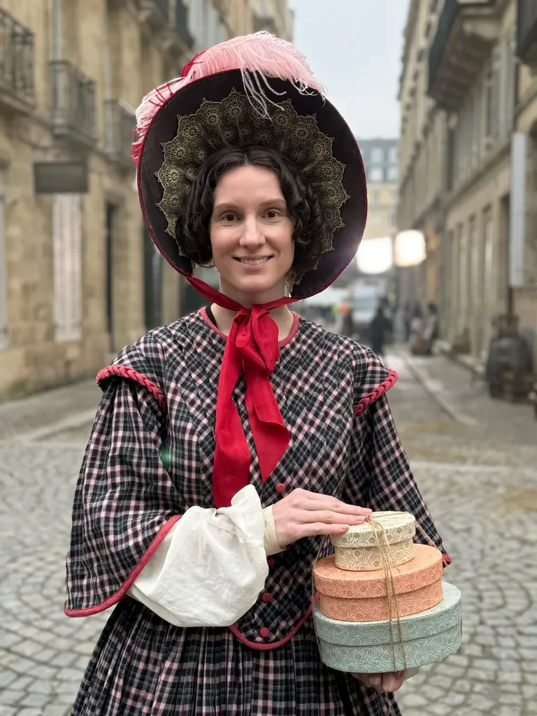 A smiling woman in a checked dress and feathered bonnet, red ribbon at her neck, holding a stack of hatboxes, colour.