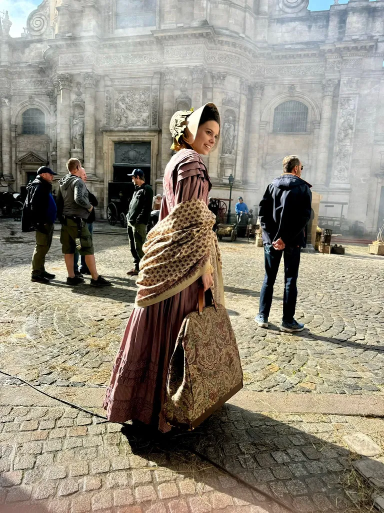 A woman in a period dress and shawl looks back in front of a carved church front, the crew around her, colour.