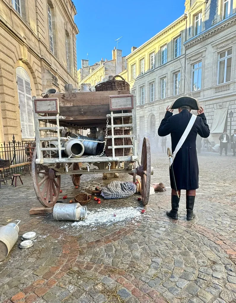 An overturned cart and milk churns spilled on the cobbles, a man in a bicorne and long coat beside it, blue sky, colour.