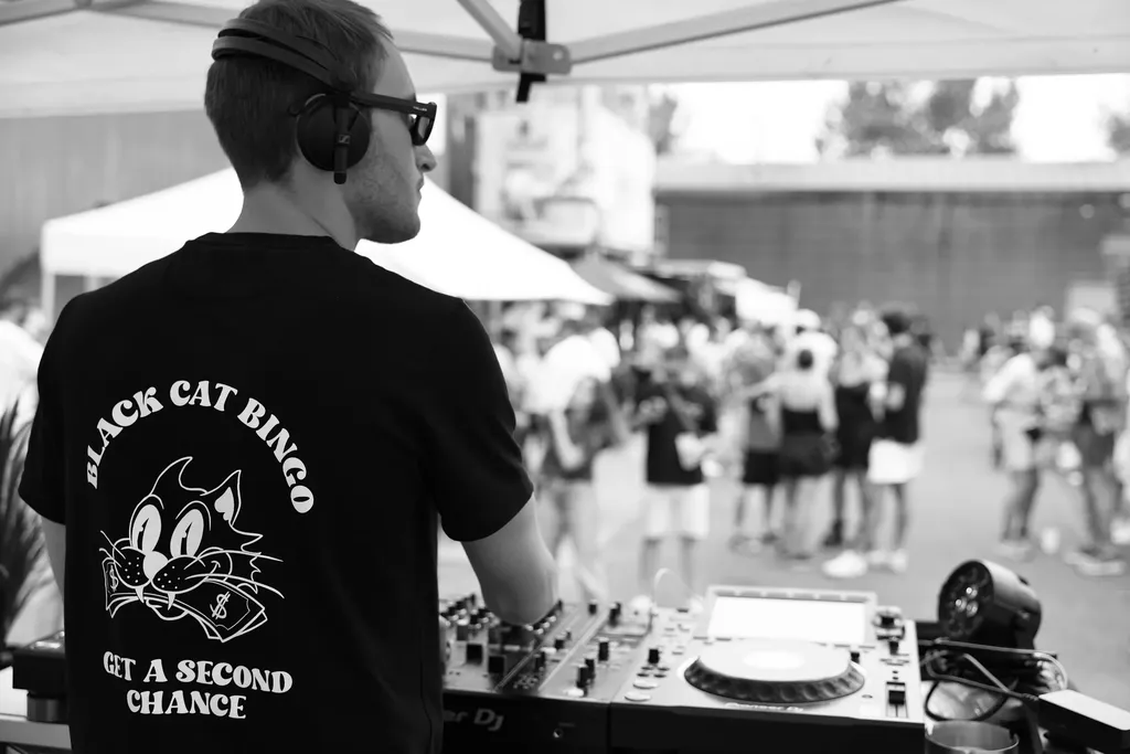 From behind, a DJ in headphones at the decks under a tent, facing an outdoor crowd, black and white.
