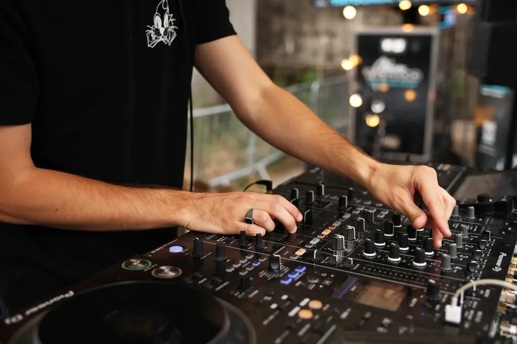 A DJ’s hands on a mixing desk, blurred string lights behind, colour.