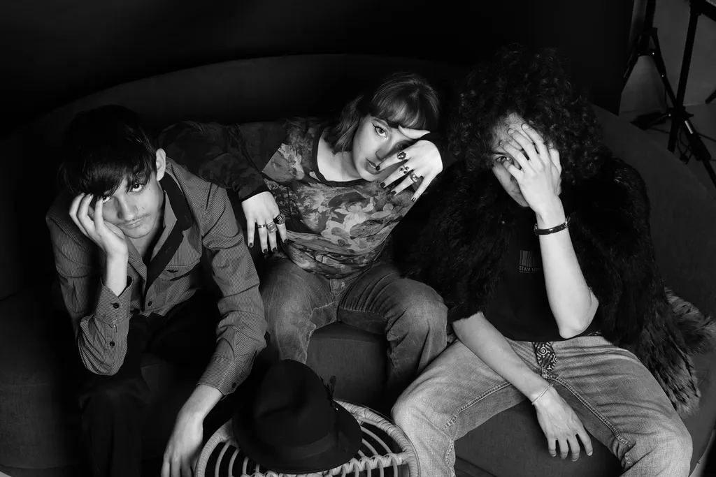 Three young people sprawled on a round sofa looking into the lens, a hat on the floor, black and white.