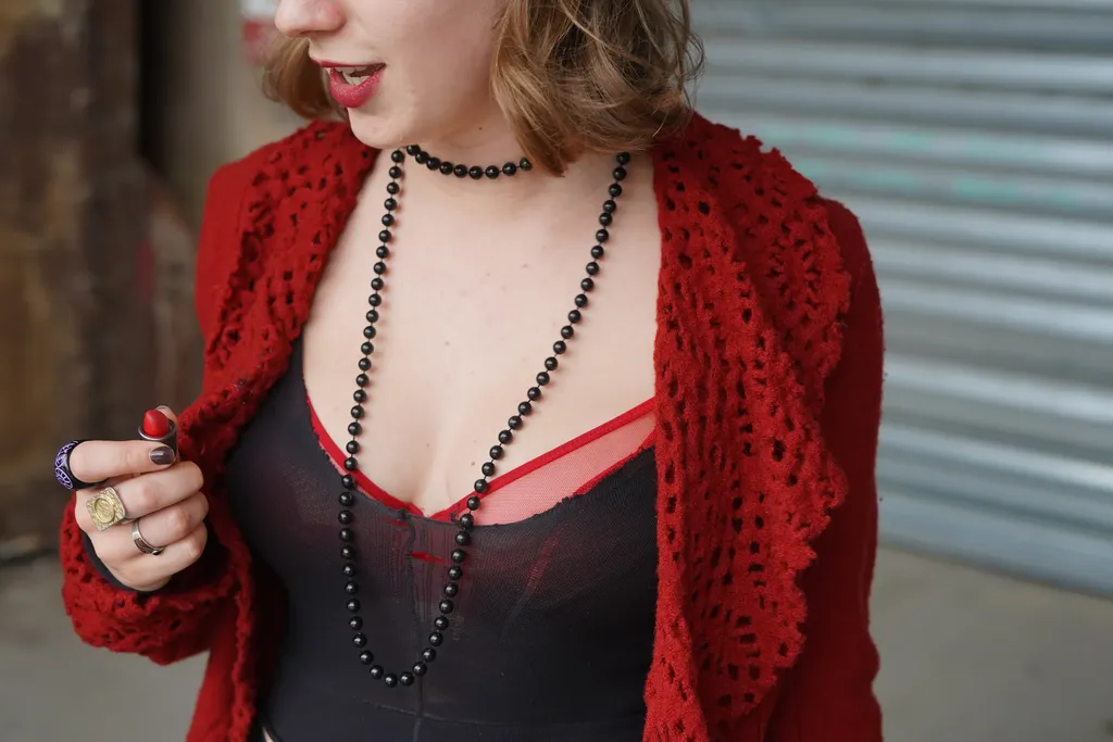 Close-up of a woman in a red crochet cardigan, black top, bead necklaces and rings, red lips, colour.