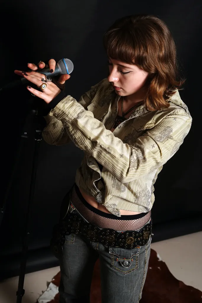 A young woman in a silver shirt holds a microphone in front of a black backdrop, colour.